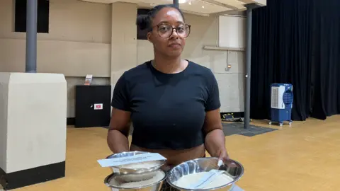 Lexi Walker looks at the camera and holds three bowls of rice in her hands. She is wearing a black t-shirt and standing in a room with a wooden floor and long black curtains hanging down one wall in the background.
