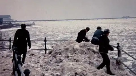 John Clague Four people stand at the sea railings among mounds of snow and ice. The sea in the background is also covered in ice. 