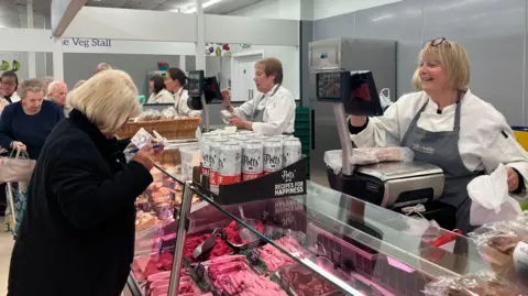 A customer buys produce at a butcher's counter. A range of sausages, mince and other meat is visible inside a glass cabinet. A woman stands behind the counter wearing a white top and blue butcher's apron - she is weighing meat on a scale.