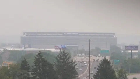 Smoke from Canadian wildfires shrouds MetLife Stadium, host of the 2026 FIFA World Cup final, as hazy orange skies reduce visibility over a busy highway in New Jersey.