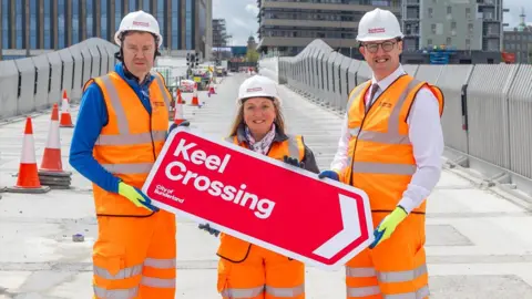 Councillor Kevin Johnston, councillor Lindsey Leonard and councillor Michael Mordey hold a red finger sign with white edging reading Keel Crossing. They are at the new footbridge. They are wearing orange protective clothing and white helmets. The grey concrete structure is still under construction. There are more new building on its other side, including the City Hall. Keel Square is also visible in the distance.