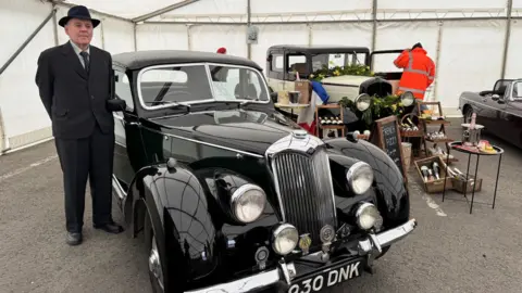 A black vintage car inside a tent, with a man in a black suit and tie and black hat stood next to it, smiling. In the background is another black and cream vintage car.