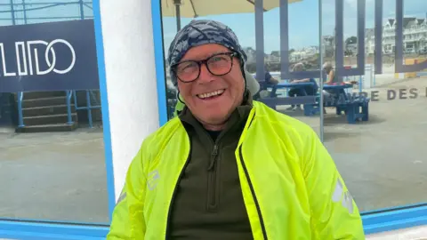 Gerry Leonard in a bright yellow jacket and a patterned head covering sits in front of a glass window with the words "LIDO" and "HAVRE DES PAS" printed on it. Behind the glass, there are blue tables with people seated and a large umbrella. Reflections in the window show nearby buildings and possibly a waterfront area.