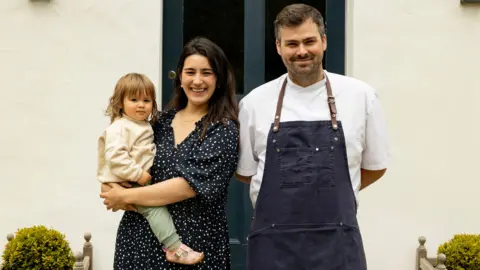 Matilda and Tom Tsappis standing outside Killiecrankie House. Matilda has shoulder length dark brown hair and is wearing a black dress with white spots and is holding a toddler on her hip. Tom has short brown hair and a trimmed beard. He is wearing a white chef's jacket and a navy blue apron with leather neck strap.