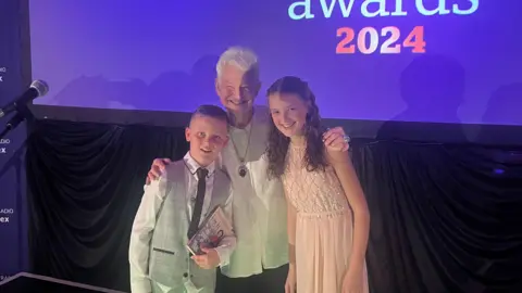 Dame Jacqueline Wilson with two young fans Hayden and Sennen Calver on the stage with a purple backdrop