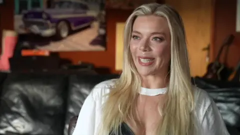 A woman with blonde hair and wearing a white cardigan, sits on a sofa.