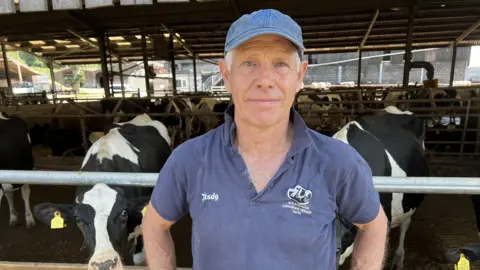 Peter Appleton, wearing a hat and polo shirt and looking at the camera. A barn full of black and white cows is seen behind him.