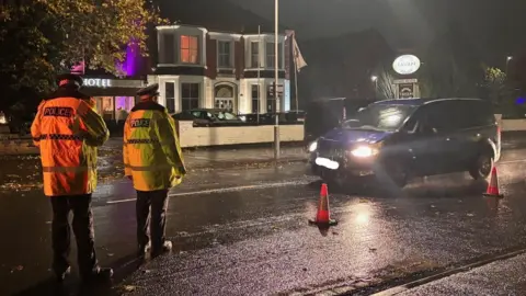Cambridgeshire Constabulary Two police officers, men, wearing green police jackets, standing on the road, stopping cars, with orange cones placed on the side of the road.