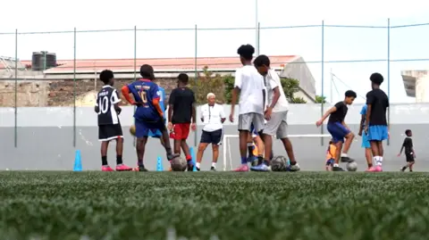 Children play football on an artificial pitch in Cape Verde while a coach oversees their progress