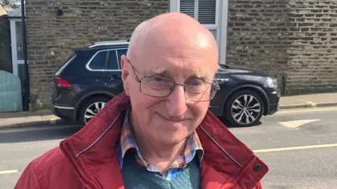 Grace Wood/BBC A man with a bald head and rectangular glasses. He is standing on the street wearing a colourful shirt, blue jumper and red coat.