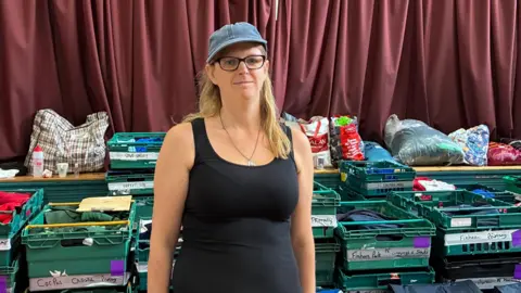 Zia Lee stands in a black vest top with a denim cap on and black glasses and long blonde hair. Behind her are green crates with school names and carefully folded uniforms in them.