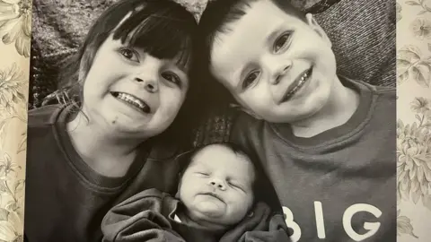 A black and white photo of a boy and girl smiling with a baby cuddled between them. The photo is hung on a wallpapered wall