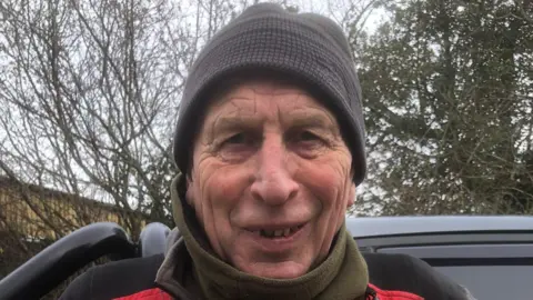 Peter Moss is looking directly into the camera and is smiling. He is wearing a grey hat, green fleece and a black overcoat. There is a grey vehicle behind him.