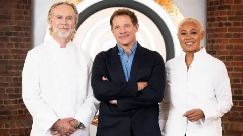 BBC/Shine TV The three judges of series 18 of MasterChef The Professionals on the set of the show. Marcus Wareing and Monica Galetti wear chefs coats and black trousers, with Matt Tebbutt standing between them, wearing a navy suit with blue shirt. Wareing has long blond hair combed back, blue eyes and a trimmed white beard. Waring has short brown hair and blue eyes. Galetti has blond short hair. The three are standing on set and smiling