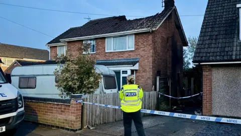 Leicester Media Online A police cordon around a house in Wigston in Leicestershire. An officer is standing facing the damaged house. The house has a large hole in the roof. There is a small caravan in the front drive. 