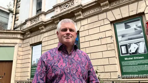 Councillor Michael Long, a grey haired man in a paisley patterned purple and pink shirt, stood outside a derelict looking stone building