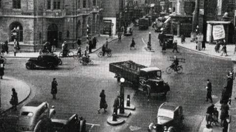 Oxford City Council  Carfax in Oxford city centre with vehicles and pedestrians. A cyclist is carrying a ladder as he rides from Cornmarket, across Carfax and to St Aldate's 