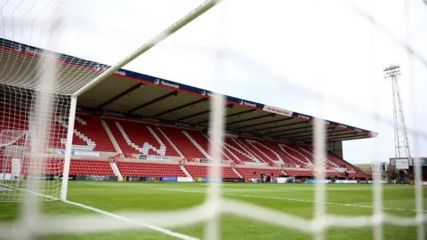 A photo of the County Ground in Swindon taken from behind the goal, with the grass pitch and red stand with the word Swindon written out in white seats.
