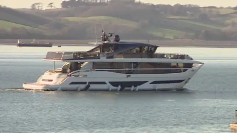 A blue and white yacht on the water with rolling green hills in the background.