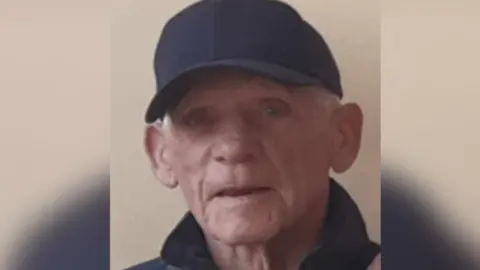 An older man with grey hair, wearing a black baseball cap and jacket.