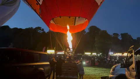 A group of people gathered around a hot air balloon basket. Their faces are lit up by a burners' flames in full force. They are surrounded by people with their phone lights, on filming the situation.