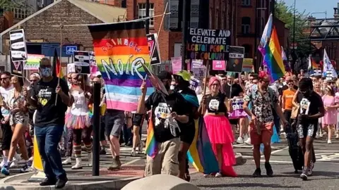 A protest march with a large crowd of people carrying banners. The most visible banner is a rainbow banner that has "Rainbow Noir" painted across it in large letters. Another visible sign says "elevate and celebrate LGBTQI people of colour". The way the photo is taken means most faces aren't visible.
