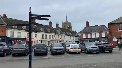 Several cars can be seen parked in bays in a town market place, with a black sign pointing to different local destinations in the foreground and a row of shops and a church in the background.