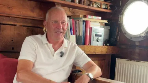 Mark Jones sits inside a traditional wooden boat cabin, leaning one arm on a wooden railing, wearing a white polo shirt. The cabin interior is lined with dark varnished wood. Behind him are shelves holding books and a large round porthole window on the right lets in bright daylight