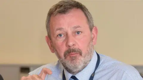 Richard Pengelly is looking into the camera. He's wearing a blue and white shirt with a tie. There's a lanyard over his neck. 