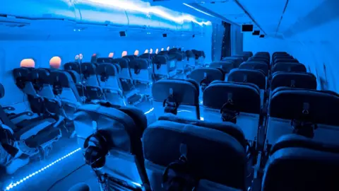 Mihaela Bodlovic What looks like the inside of a plane - aircraft seats with blue strip lighting and headphones hanging on the back of each one. On one side, there is a set-up made to look like a plane wall with windows