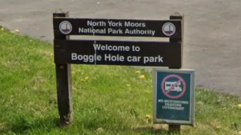 Google The picture shows a wooden sign for the North York Moors National Park Authority at the entrance to Boggle Hole car park. The sign stands on a patch of grass beside a paved area. Below it a smaller notice displays a symbol indicating no motorhome parking overnight.