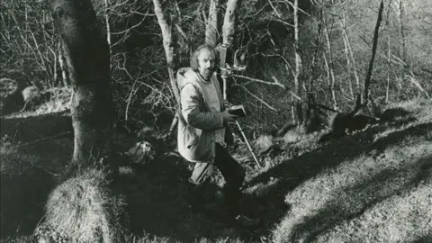 Paul Hill A black and white image of a man stood in woodlands in the 1980s holding a notebook and standing next to a camera on a tripod. Around him are several trees with grass on the ground. He wears a coat over a light jumper and has a beard.