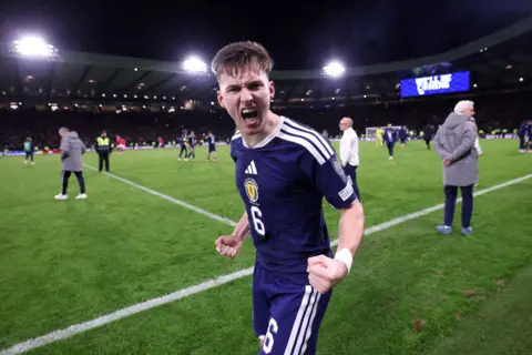 Getty Images Kieran Tierney is wearing a blue Scotland kit and his mouth is open in a shout, his fists clinched as he celebrates at the side of the pitch.
