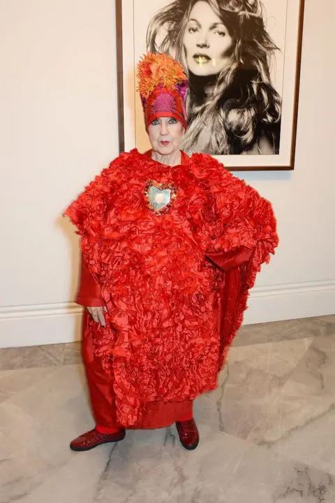 Getty Images Molly Parkin attends a private view of Bailey's Stardust, a exhibition of images by David Bailey supported by Hugo Boss, at the National Portrait Gallery on February 3, 2014 in London, England. She is stood looking at the camera and there is a framed black and white picture of Kate Moss behind her. She has her left arm on her hip and is wearing a red floral kaftan dress and a red and orange head wrap 