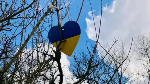 Elmbridge Borough Council A blue and white Ukrainian heart decoration hanging from a tree against a blue sky.