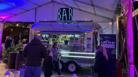 Ollie Conopo/BBC People queue at a small silver trailer being used as a bar. The trailer is underneath a white covering. 