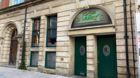 A sand coloured brick building with four green edged windows and two green doors with "R" printed on the front. The sign for the pub "Rosie O'Leary, Bar and Tap" is above the doors.