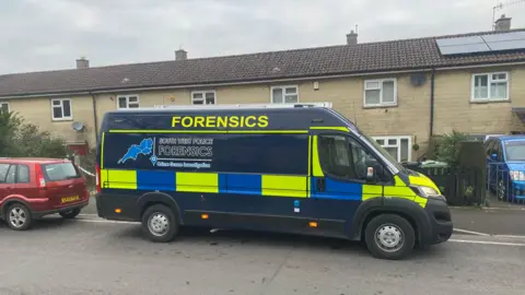 A police forensics van is parked on a road with a row of houses in the background and a red car parked on the left 