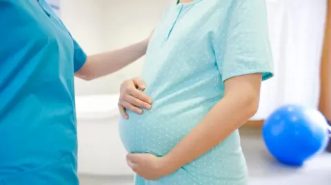 A pregnant lady standing and holding her stomach. A midwife is standing next to her. An exercise ball is in the background.