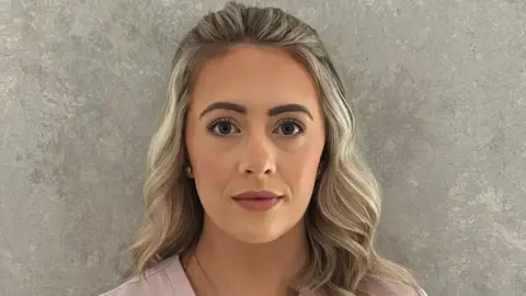 Claire Harkin has long wavy blonde hair. She has some mascara and pink lipstick on and is wearing a pink top.