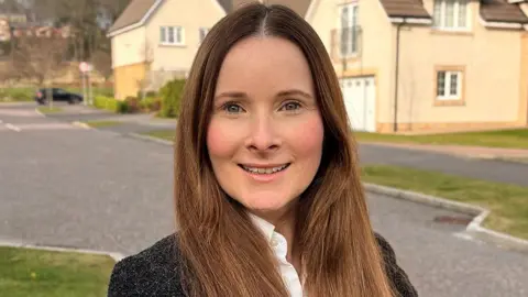 Natasha Douglas, who has long brown hair, smiles while looking directly at the camera. She is wearing a dark jacket and white shirt, with houses in the background 