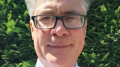 A close-up image of a man with glasses and grey hair who is wearing a shirt. He is very prominent in the photo, looking at the camera, and in front of bushes that fill the background.