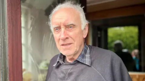A man called Eddie Amhof stands in the doorway of his home. He is an elderly man and has white hair and bushy eyebrows. He is wearing a black jumper with a black checkered collared shirt underneath.