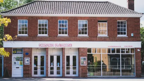 The outside of Norwich Theatre Playhouse. The building is made from bricks and has white signage with red lettering which says, NORWICH PLAYHOUSE, on the front above the white doors.