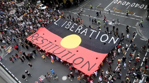 Reuters Una imagen a vista de pájaro de una manifestación, con un gran grupo de personas marchando por una calle. Llevan una gran bandera roja, negra y amarilla –la bandera aborigen australiana– con las palabras “Liberación Restitución” escritas en ella.