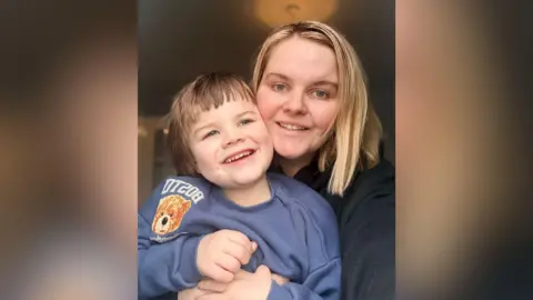 Leah McGregor A woman with shoulder length blonde hair, holding her five-year-old son. The boy has short brown hair and is wearing a blue top. They are both smiling. 