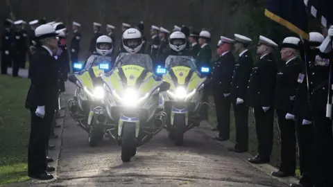 Essex Police Three police motorcycles are driven along a track at the crematorium. They are flanked by immaculately dressed officers wearing black suits and white hats and who are lining the track.