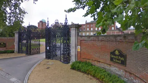 Google Red brick wall is outside of the school with a black gat which is opened. The school is made of red bricks and has a grass area outside the front.