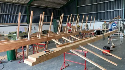 Alex Hunter Wooden sail frames can be seen propped on red metal stilts within a workshop. The frames have prongs of wood that spread horizontally off one main wooden plank.