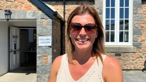 A woman with shoulder-length blonde hair in a knitted vest and sunglasses smiling in front of a polling station.